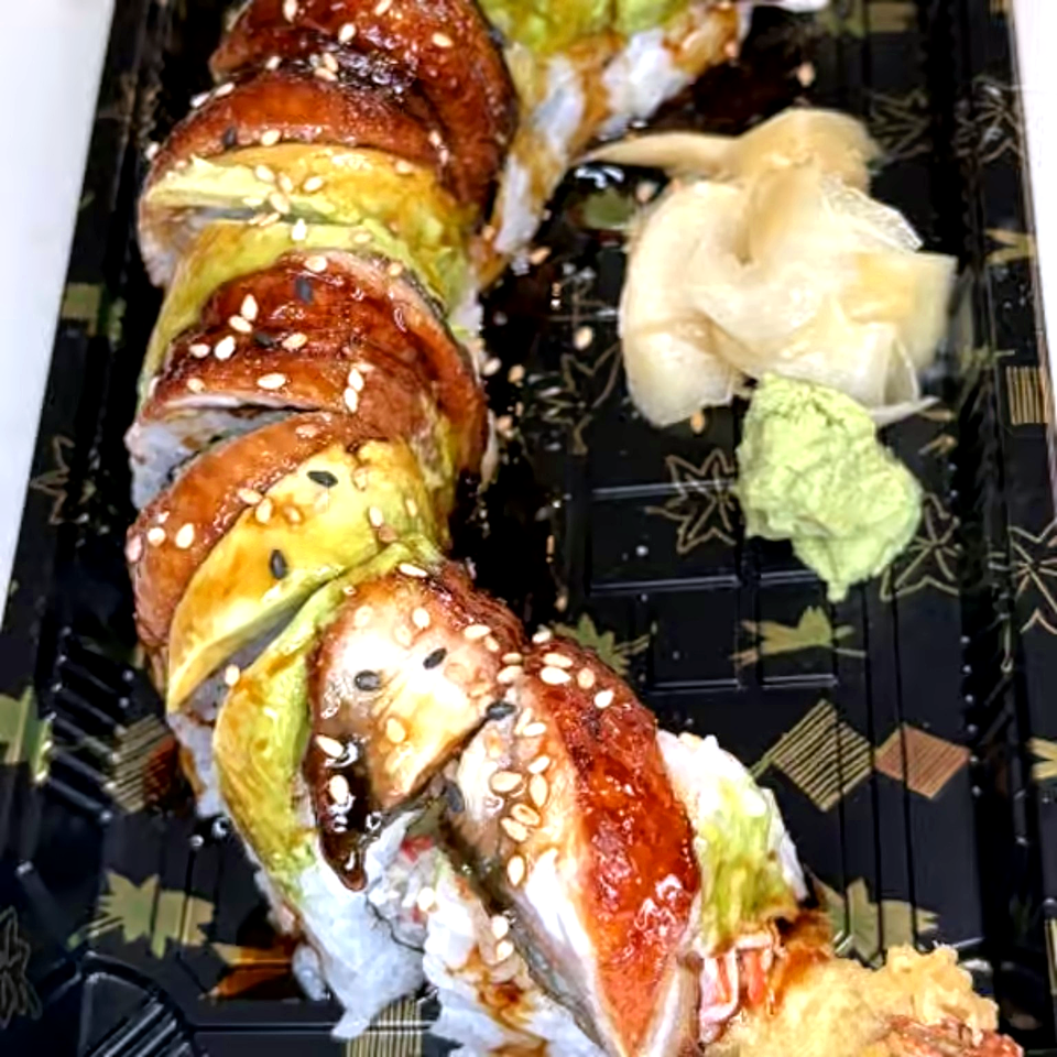 Sliced sushi roll topped with sesame seeds and avocado, served with pickled ginger and wasabi on a black patterned plate.