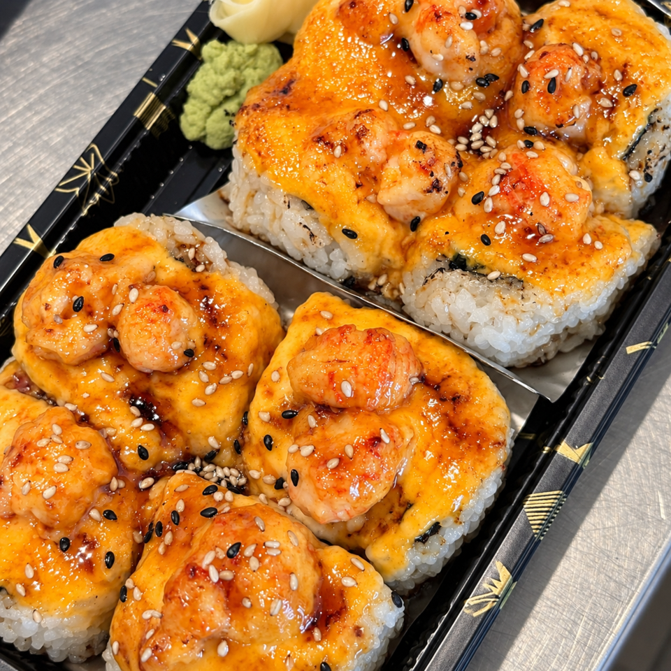 Close-up of baked sushi rolls topped with shrimp, creamy orange sauce, sesame seeds, and served with wasabi and pickled ginger.
