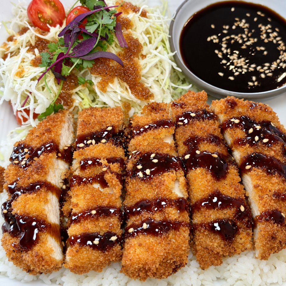 Breaded chicken katsu slices drizzled with dark sauce and sesame seeds, served with shredded cabbage, herbs, tomatoes, and rice.