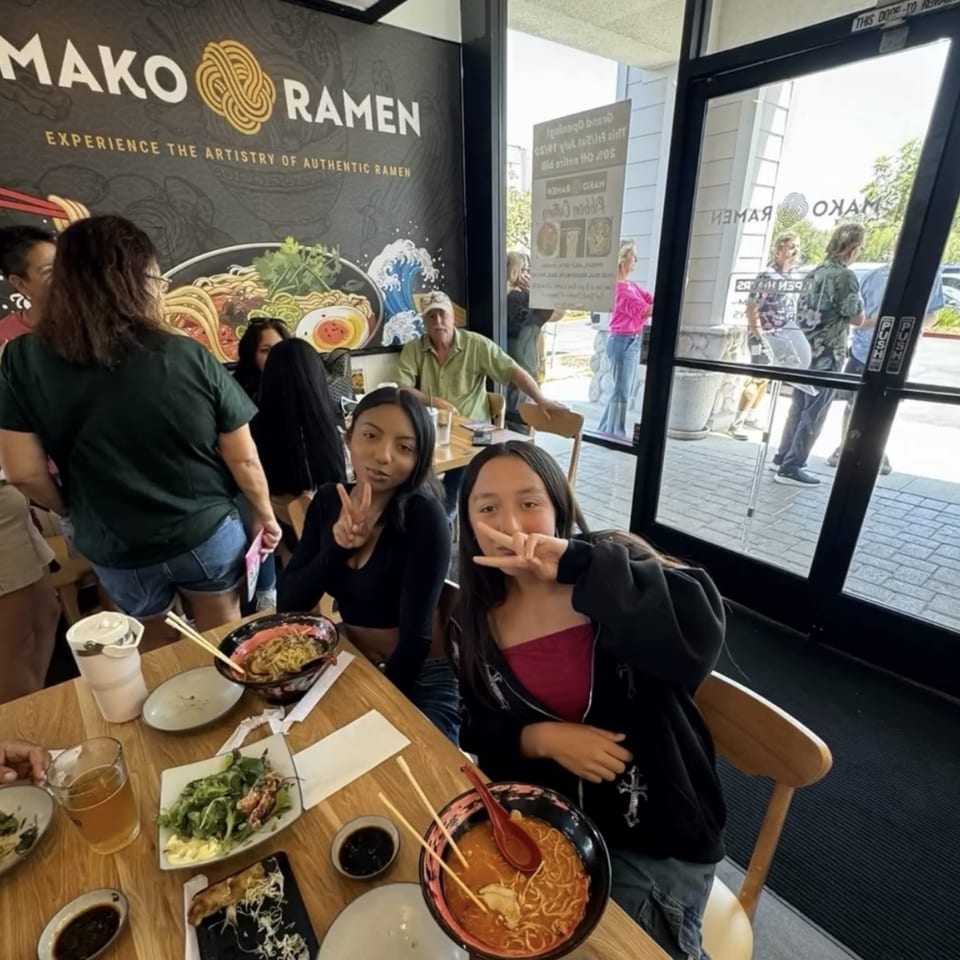 Diners enjoying ramen and other dishes at Mako Ramen, with two young women making peace signs.