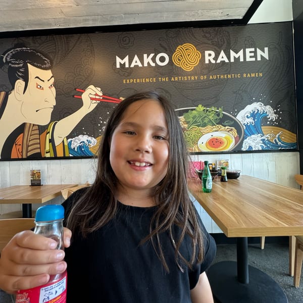 A smiling child holds a drink in front of a Mako Ramen mural with a samurai and ramen bowl.