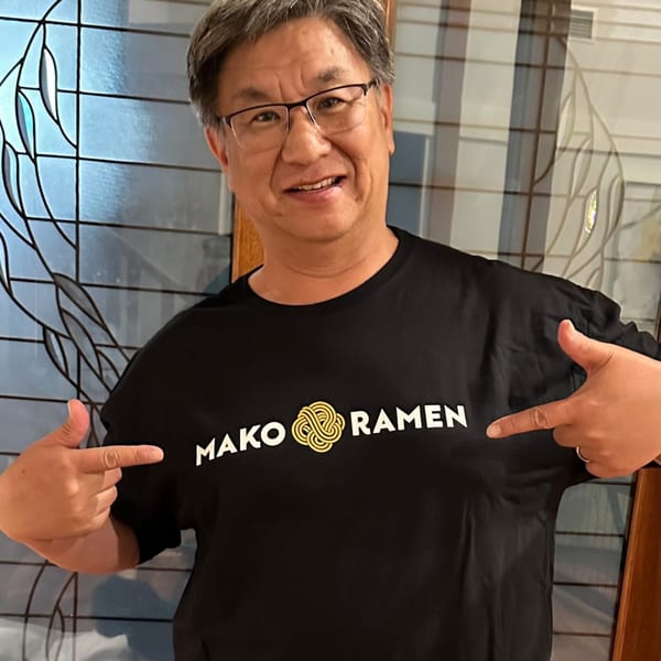 A smiling man in glasses points to the Mako Ramen logo on his black t-shirt.