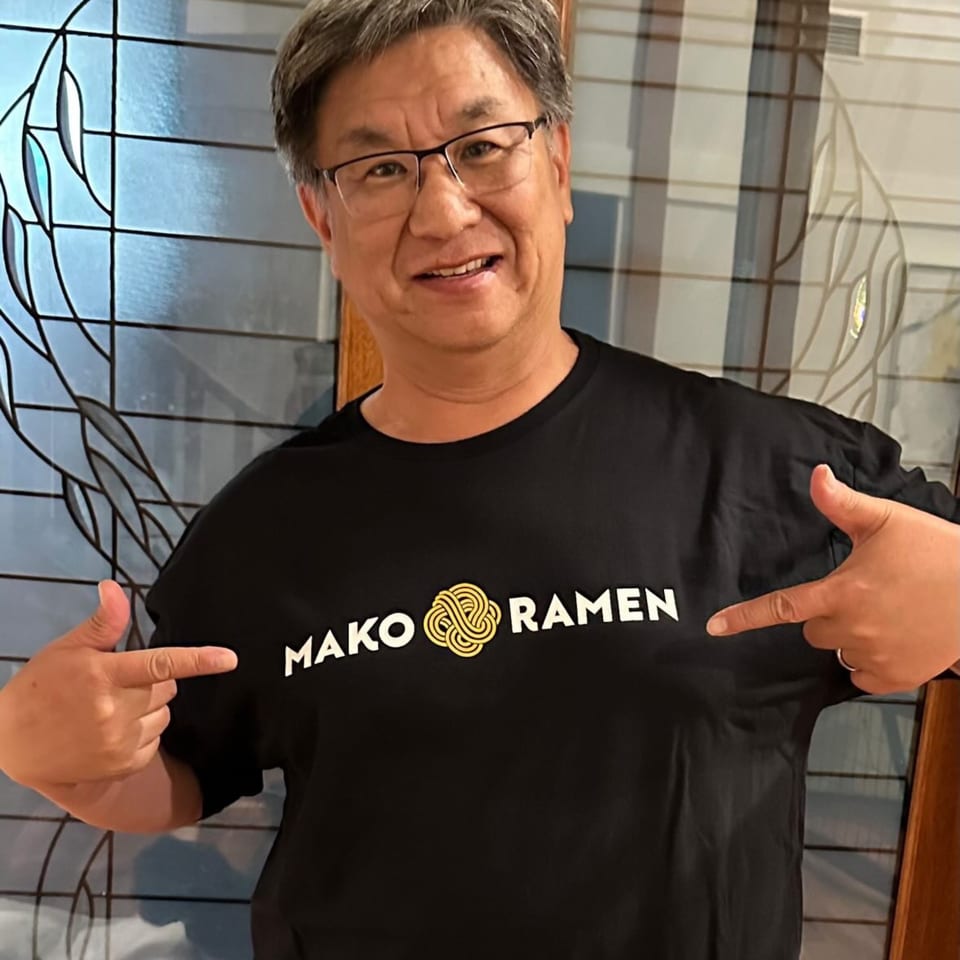 A smiling man in glasses points to the Mako Ramen logo on his black t-shirt.
