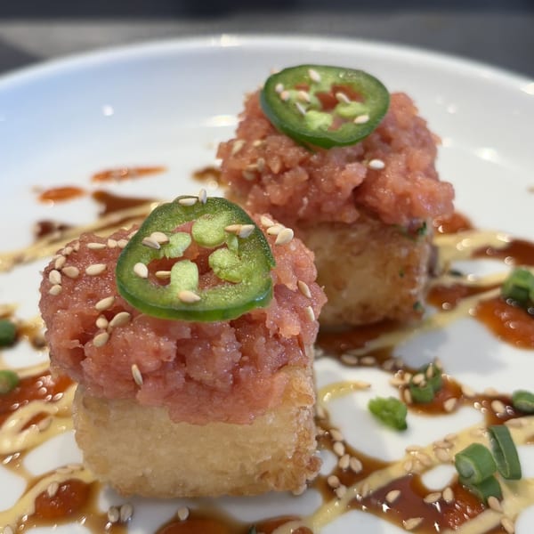 Two crispy rice cakes topped with spicy tuna, sesame seeds, and a jalapeño slice on a white plate.