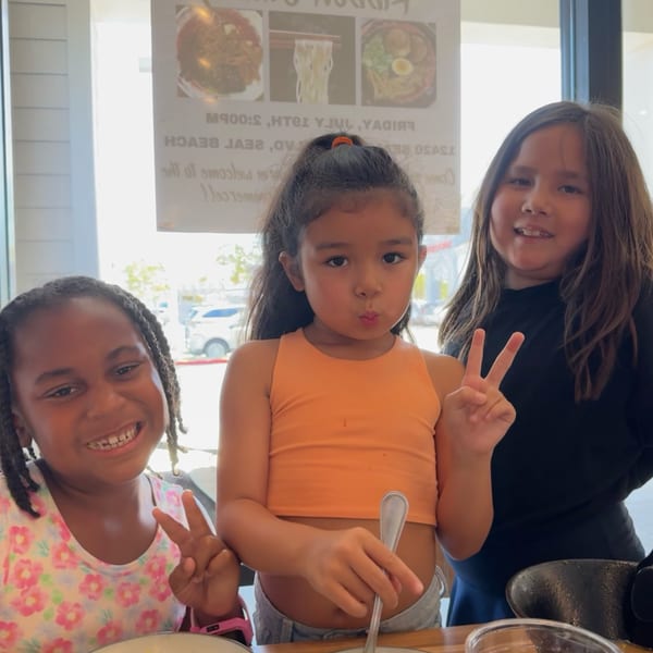 Three young girls smiling and making peace signs at Mako Ramen, with menu posters visible.
