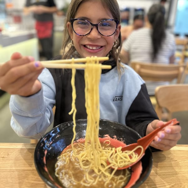 A smiling girl in glasses holds up a long strand of Mako Ramen noodles with chopsticks.