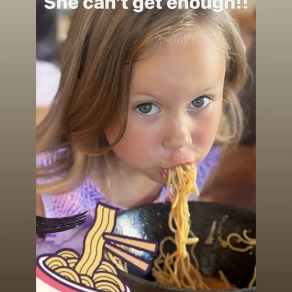 A young girl with blonde hair slurps ramen noodles from a black bowl at Mako Ramen.