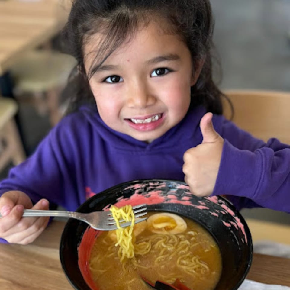 A happy child in a purple hoodie gives a thumbs up while eating Mako Ramen.