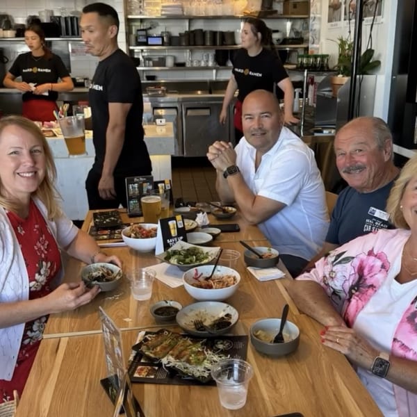 Four happy diners at Mako Ramen with bowls of ramen and appetizers on a wooden table.