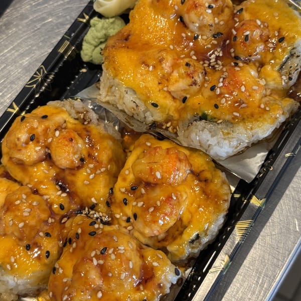 Close-up of baked sushi rolls topped with shrimp, melted cheese, and sesame seeds in a black tray.