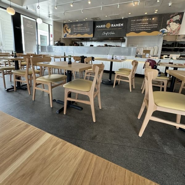 Interior of Mako Ramen, showing light wood tables and chairs, with the kitchen and branding in the background.