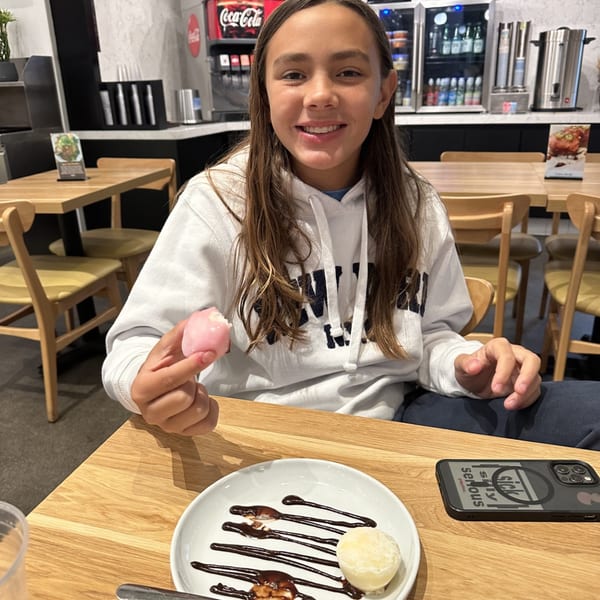 A smiling girl holds a pink mochi, with another mochi and chocolate sauce on a plate.